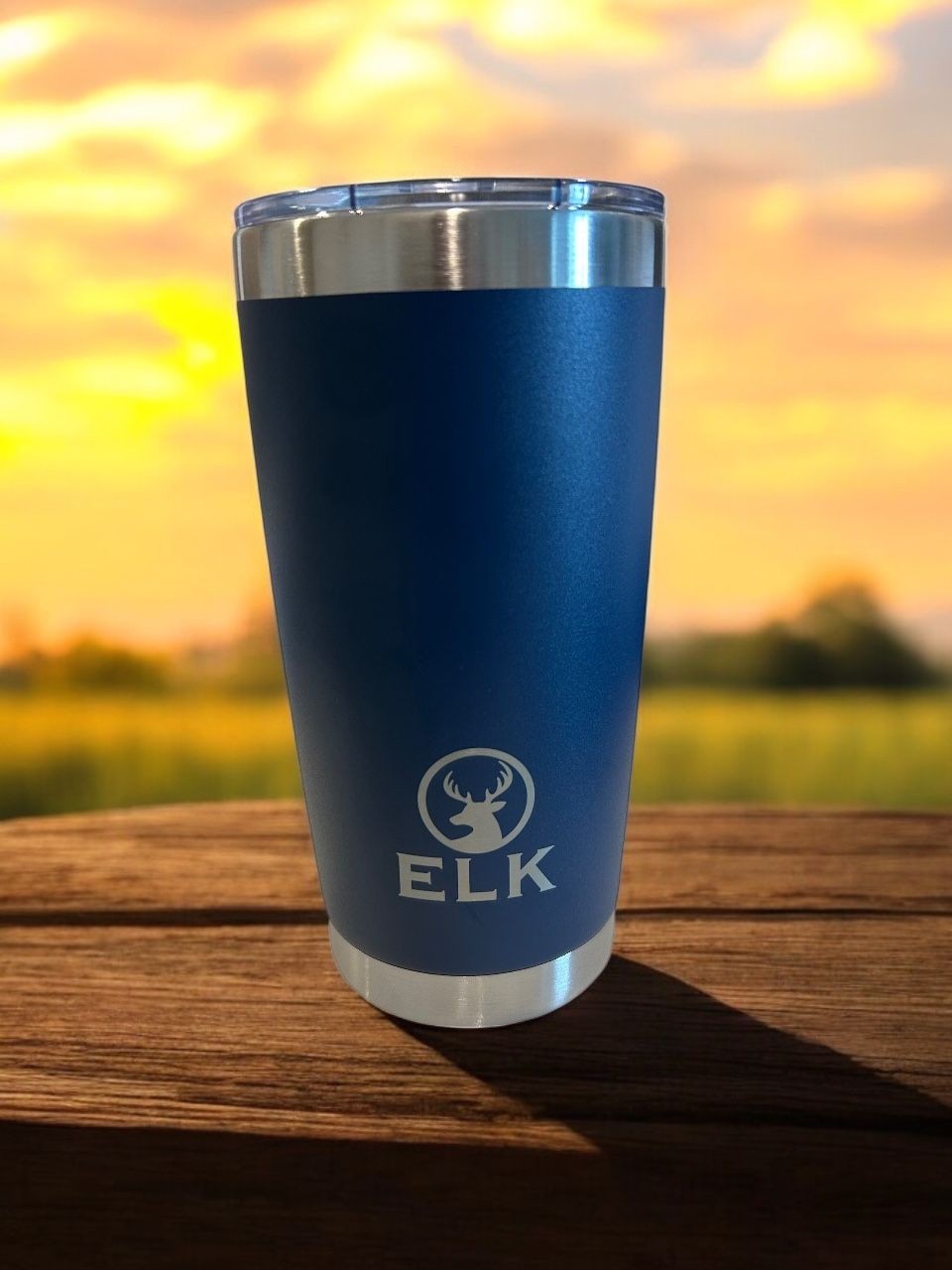 Blue travel tumbler with elk logo on a wooden table, sunset in the background.